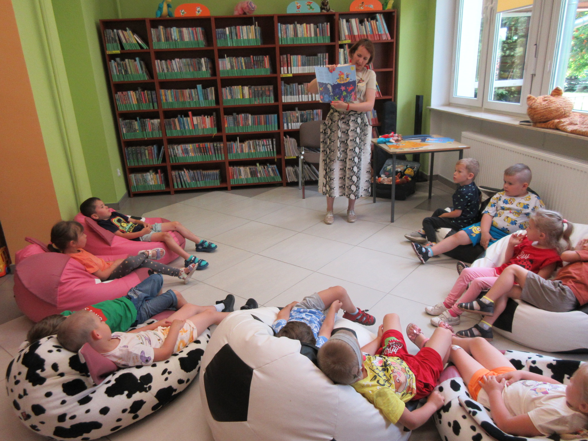 Spotkanie autorskie z Katarzyna Czubała Vyborow. Autorka stoi z rozłożoną książką z ilustracjami. Dzieci leżą na podłodze w półokręgu.